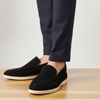 Black loafers worn with navy pants on a light wooden floor.