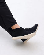 Black slip-on shoes with white soles worn by a person on a light gray background