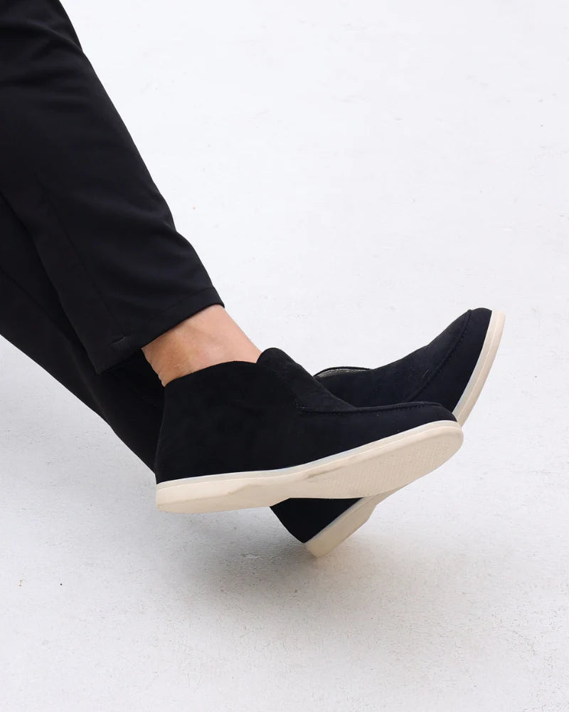 Black slip-on shoes with white soles worn by a person on a light gray background
