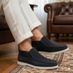 Person wearing black slip-on shoes with white soles in a living room setting.