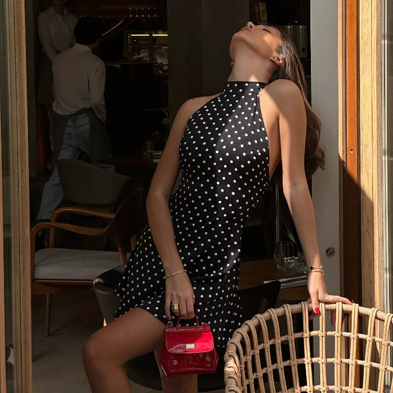 Woman in a black polka dot dress holding a red handbag, standing in a room with chairs and a door.