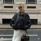 Woman wearing a black leather jacket and sunglasses, standing on a street with a building in the background.