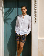 Man wearing a white shirt and patterned shorts standing against a blue wall.