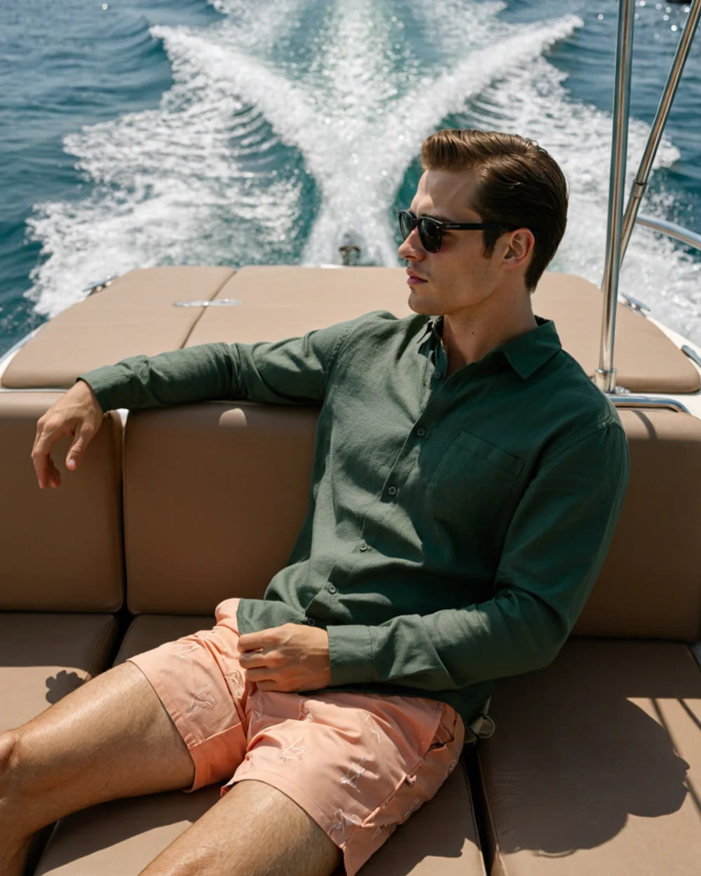 Man sitting on a boat wearing sunglasses and a green shirt with peach shorts, with water and wake in the background.
