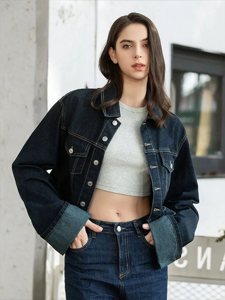 Woman wearing a denim jacket and jeans standing against a light-colored wall.