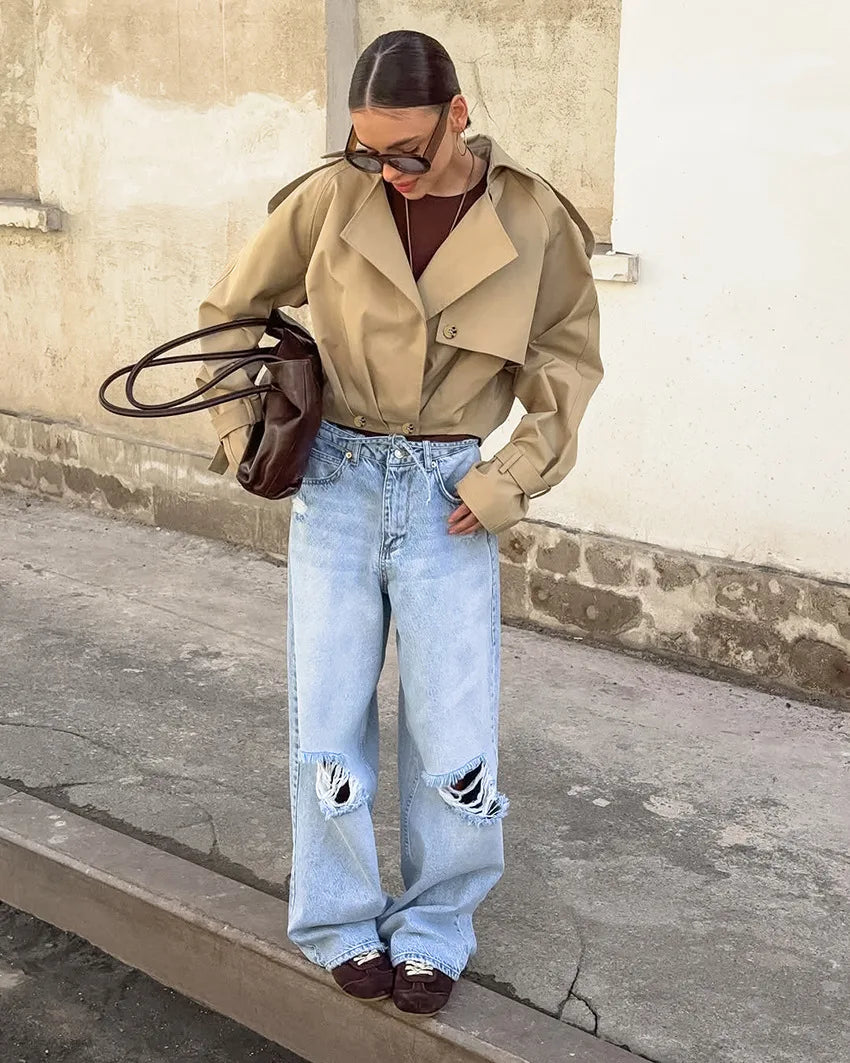 Person wearing a beige trench coat and light blue jeans standing against a wall.