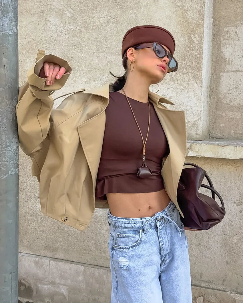 Woman in a stylish outfit with a beige coat, brown top, and blue jeans against a stone wall.