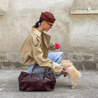 Person in beige coat and brown hat with a small dog on a leash against a concrete wall.