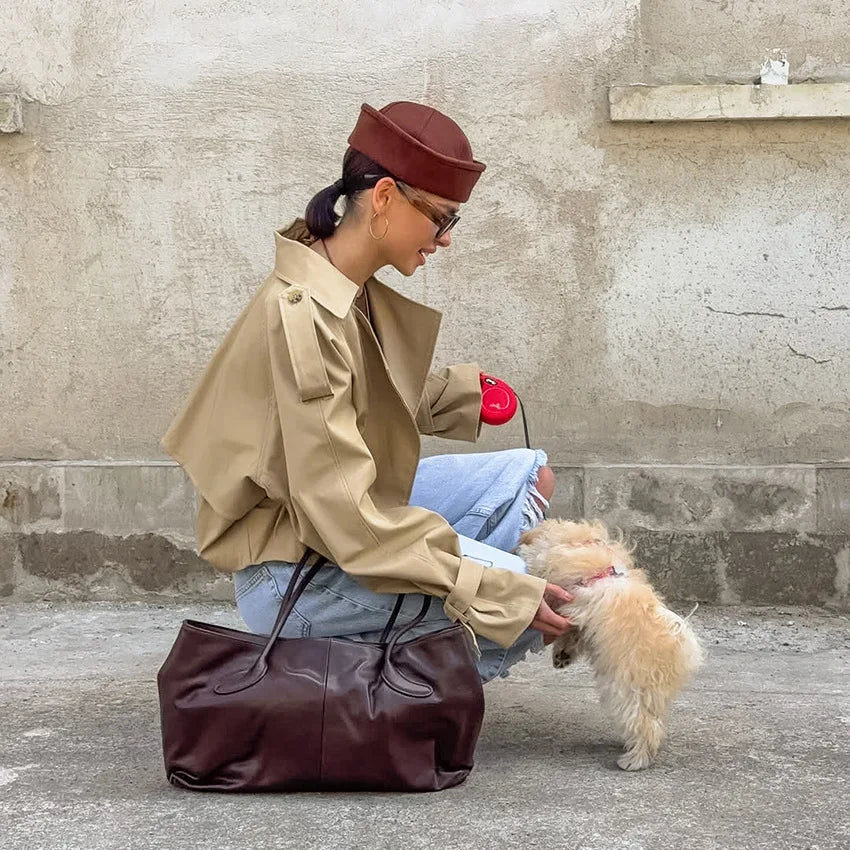 Person in beige coat and brown hat with a small dog on a leash against a concrete wall.