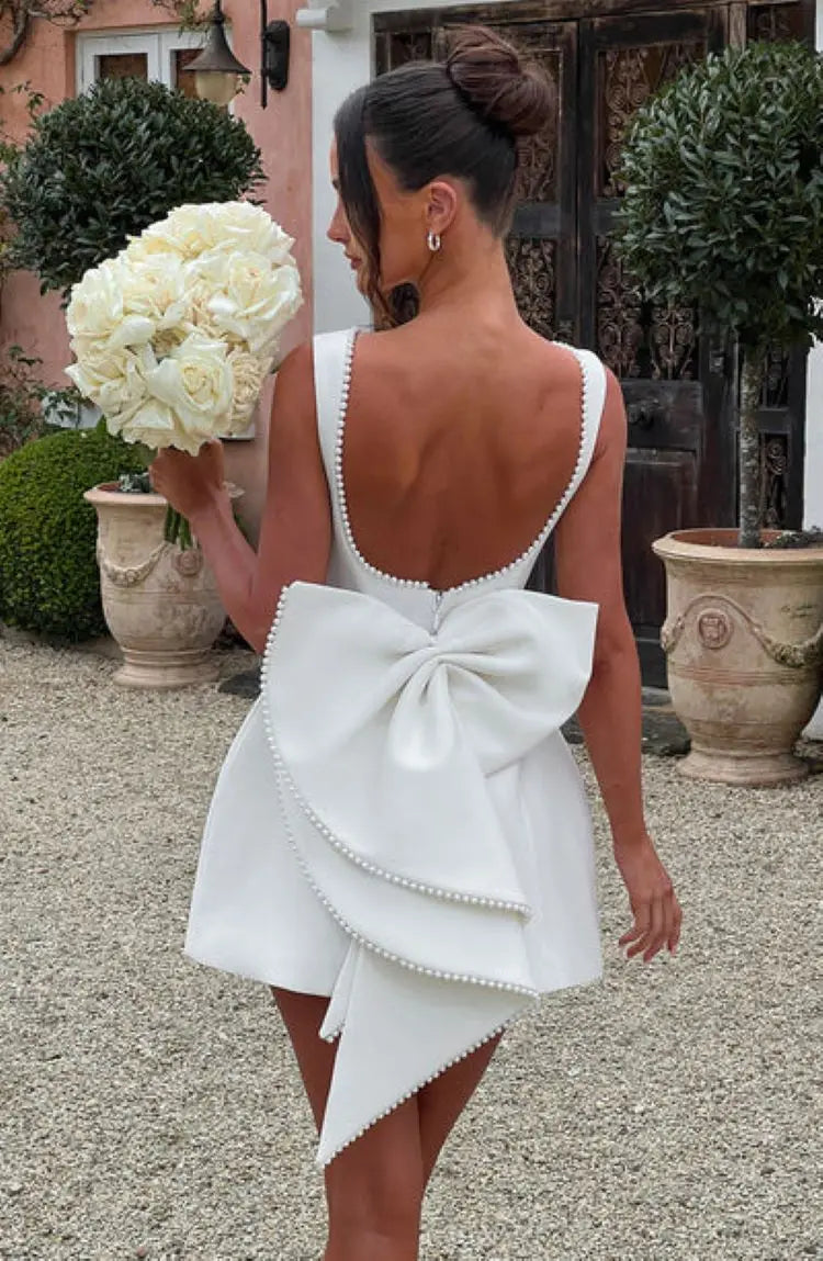Woman in a white dress with a large bow holding a bouquet of white flowers outdoors.