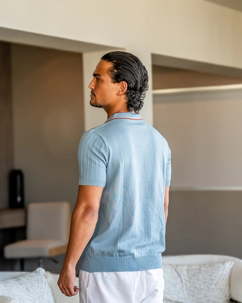 Man wearing a light blue sweater and white pants standing in a room with neutral decor.