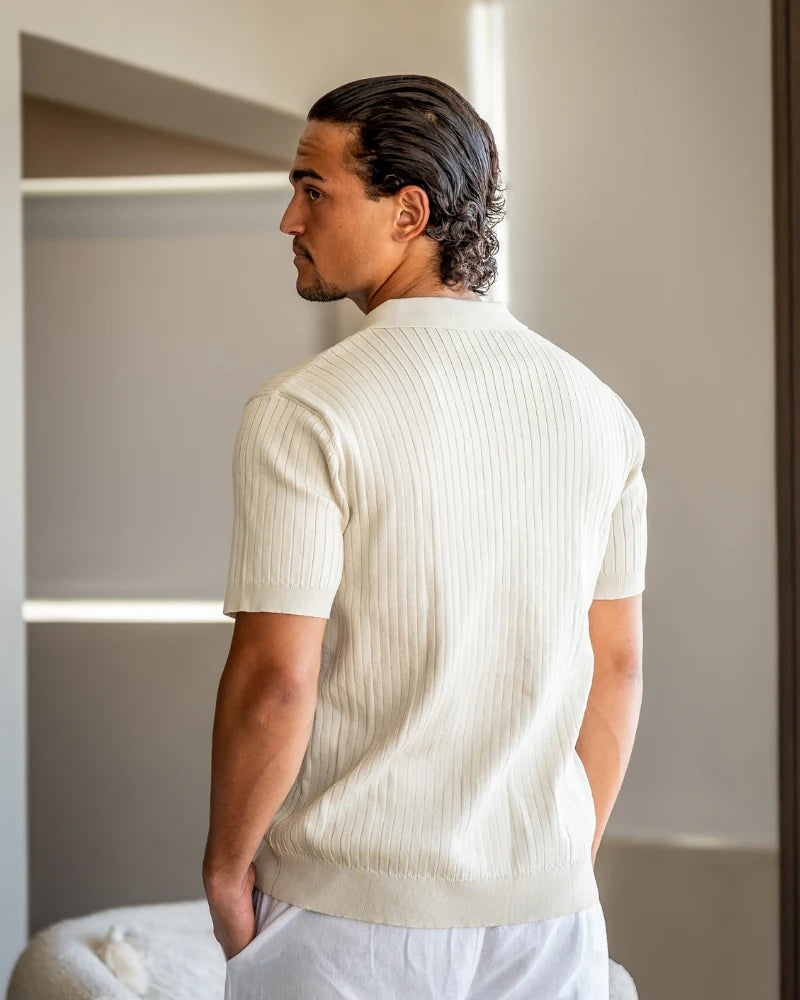 Man wearing a white ribbed sweater and white pants in a minimal indoor setting.