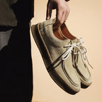 Person holding a pair of green suede shoes with white laces against a beige background