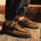Brown suede shoes worn with dark pants on a wooden floor.