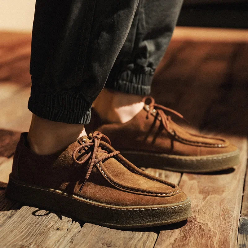 Brown suede shoes worn with dark pants on a wooden floor.