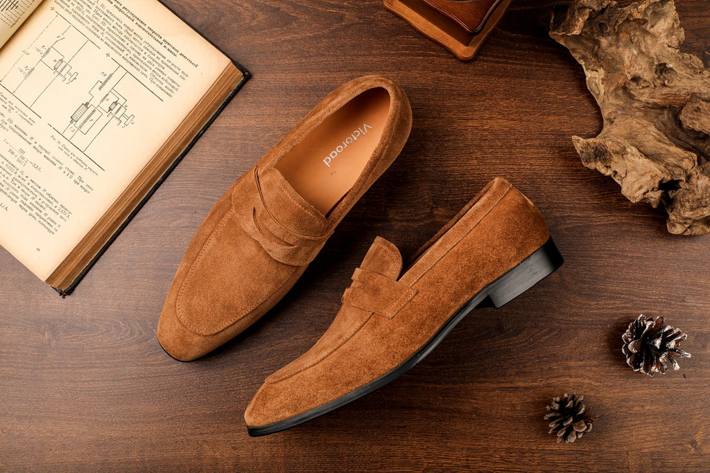 Brown suede loafers on a wooden surface with an open book and decorative elements.