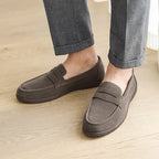 Brown loafers worn with rolled-up gray pants on a wooden floor.