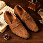 Brown suede loafers on a wooden surface with an open book and decorative items.
