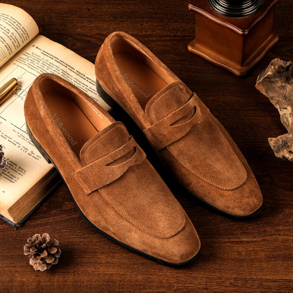 Brown suede loafers on a wooden surface with an open book and decorative items.