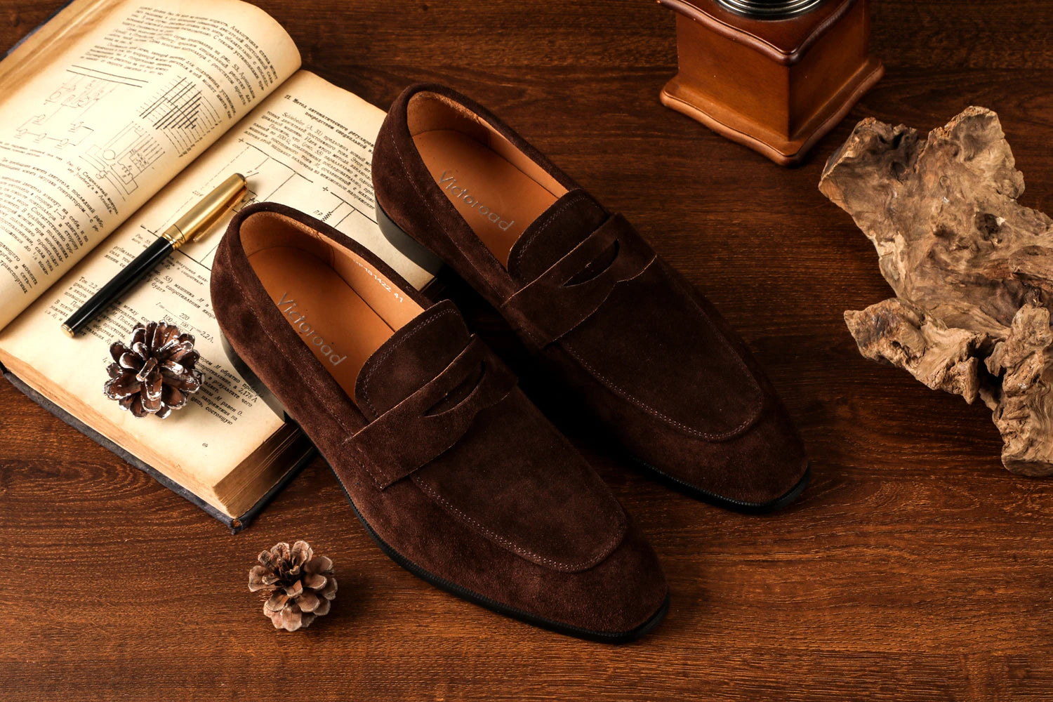 Brown suede loafers on a wooden surface with an open book and decorative items.