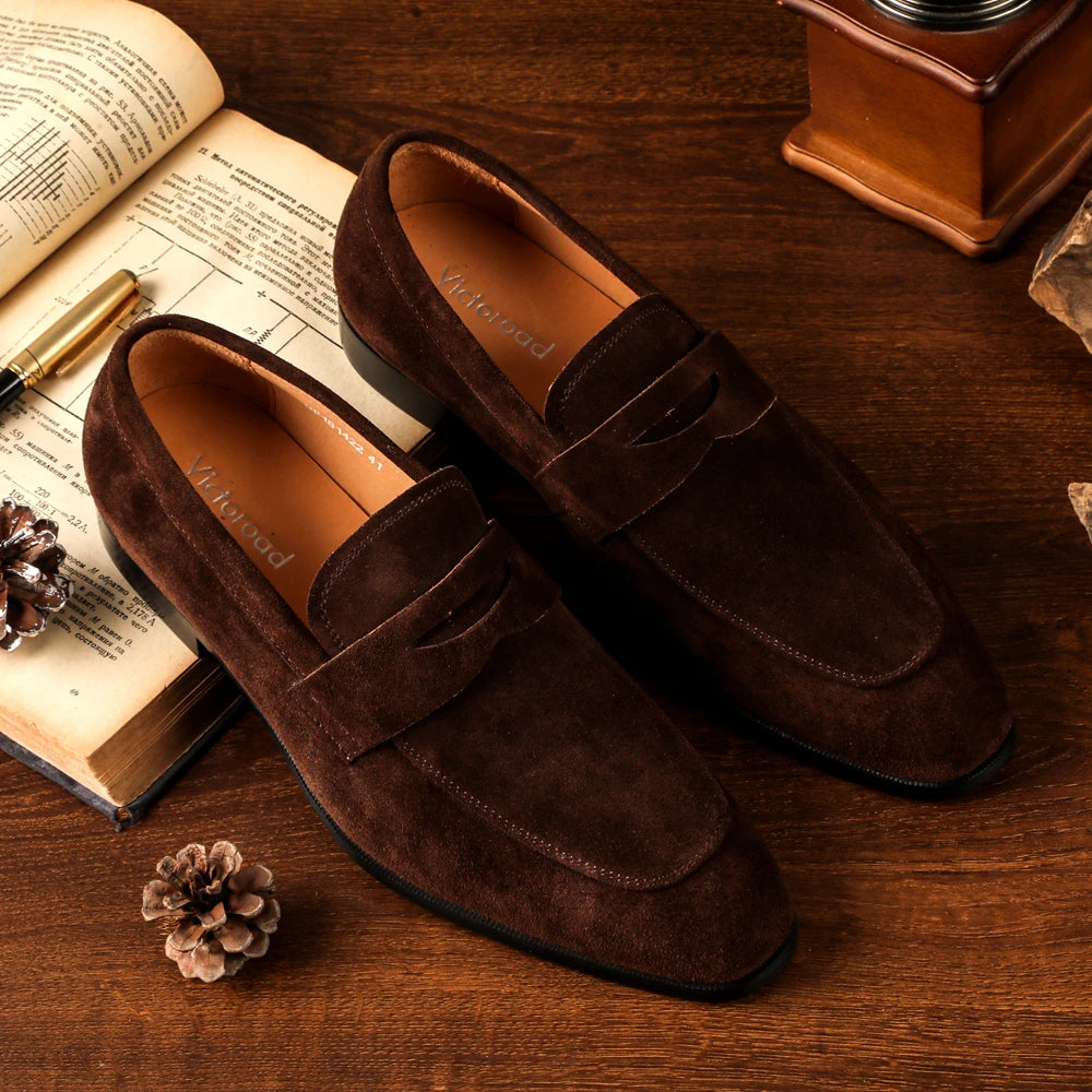 Brown suede loafers on a wooden surface with an open book and decorative items.