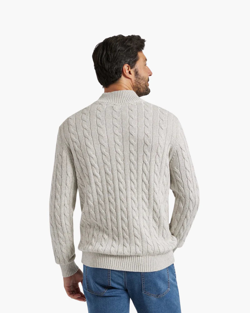 Man wearing a light gray cable knit sweater and blue jeans on a white background