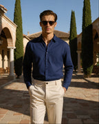 Man wearing a blue shirt and sunglasses in an outdoor setting with buildings and trees.