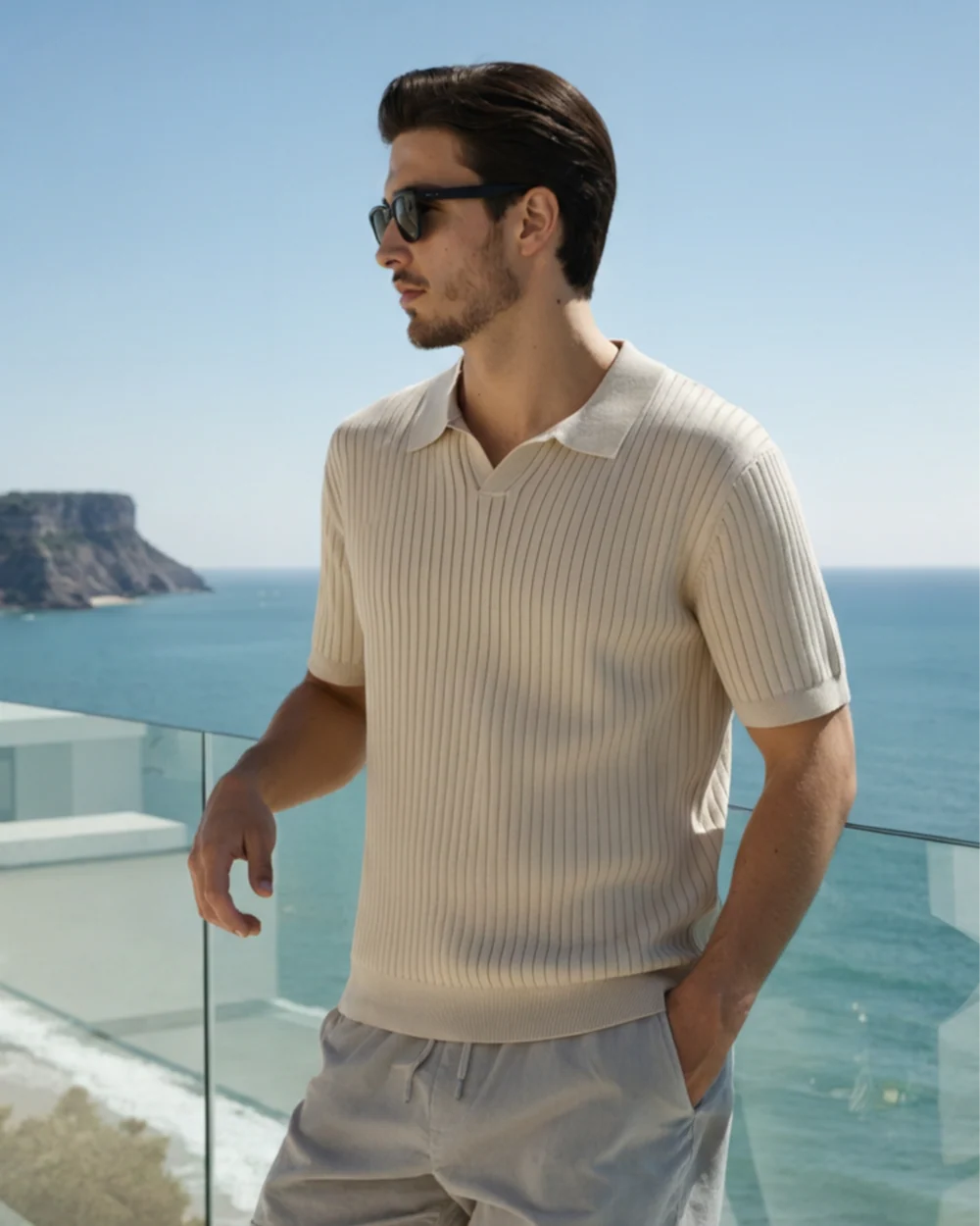 Man wearing sunglasses and a beige ribbed sweater with a scenic ocean view in the background