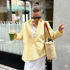 Woman in yellow jacket holding a drink and a woven bag on a city street.